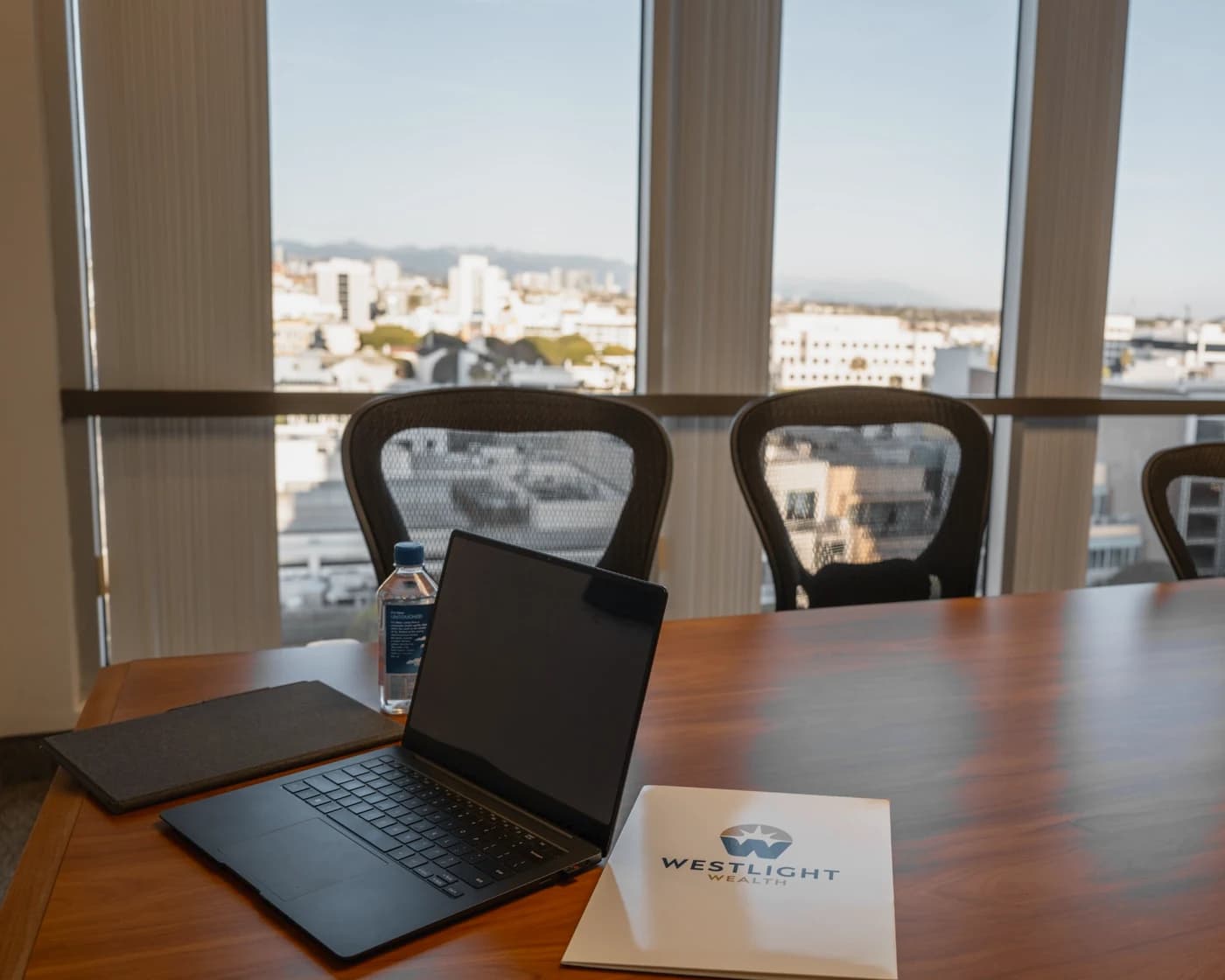 The meeting room at 100 Wilshire Blvd, Suite 700, Santa Monica: a wood table set with a laptop and a Westlight Wealth folder, mesh chairs, and tall windows over the city with the mountains beyond.